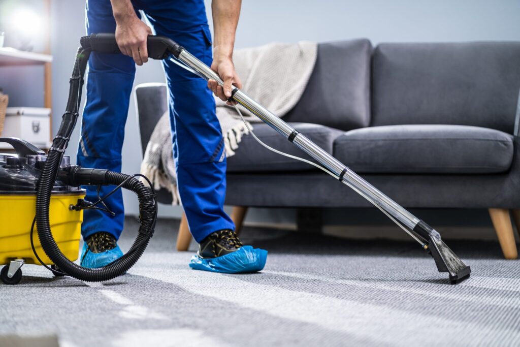 Teppichreinigung Berlin &ndash; Mitarbeiter saugt Teppich mit Nasssauger vor grauem Sofa